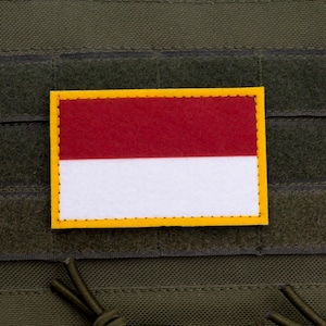 Op de afbeelding: Een rechthoekige patch met de Indonesische vlag, met een rode streep boven een witte streep. De vlag is omrand door een gele rand en is bevestigd op een groene stoffen achtergrond.