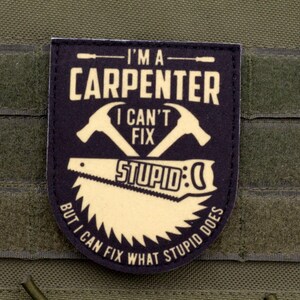 Può includere: Toppa blu navy con la scritta "I'M A CARPENTER I CAN'T FIX STUPID:)" e immagini di un martello e una sega. La toppa è su uno sfondo di tessuto verde.