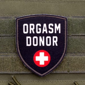 Pode incluir: Um emblema tático preto em forma de escudo com o texto branco ORGASM DONOR acima de um círculo vermelho contendo uma cruz branca. O emblema está fixado em uma superfície de tecido tático verde-oliva com tiras horizontais para equipamento.