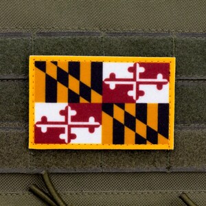 Puede incluir: Un parche rectangular con el diseño de la bandera de Maryland, con borde amarillo. La bandera muestra un patrón de cuadros negros y dorados, cruces blancas y granates, sobre un fondo verde oliva.