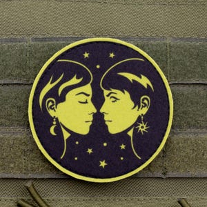 Puede incluir: Un parche redondo con dos caras de perfil amarillas, enfrentadas, sobre un fondo azul oscuro con estrellas. El parche tiene pendientes decorativos y está colocado sobre una tela verde.