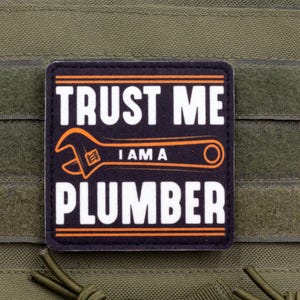 Può includere: Una toppa rettangolare nera e arancione con la scritta "TRUST ME I AM A PLUMBER" e una grafica di chiave inglese. La toppa è attaccata a uno sfondo di tessuto verde.