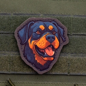 May include: A patch featuring a Rottweiler dog's head with a smiling expression. The dog has black fur with orange and brown markings. The patch is attached to a green fabric background with hook and loop fasteners.