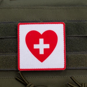 Puede incluir: Un parche blanco con un corazón rojo y una cruz blanca en el centro. El parche tiene un borde rojo y está unido a un chaleco táctico verde.