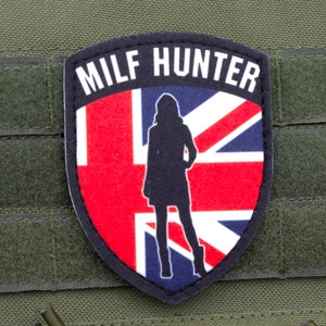 May include: A patch with the text "MILF HUNTER" above a silhouette of a woman in front of a Union Jack flag. The patch is black with a red, white, and blue design. The patch is attached to a green fabric.