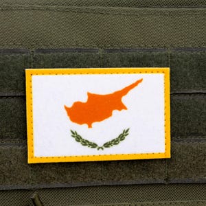 May include: A rectangular patch featuring the flag of Cyprus, with a white background, an orange island silhouette, and olive branches. The patch has a yellow border and is affixed to a green fabric surface.