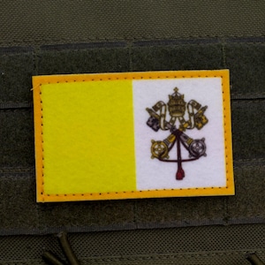Può includere: Una toppa gialla e bianca con lo stemma della Città del Vaticano. La toppa è cucita su un giubbotto tattico verde.
