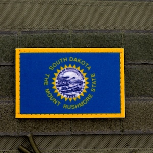 May include: A rectangular patch featuring the South Dakota state flag. The flag is blue with a central seal surrounded by the words "South Dakota" and "Mount Rushmore State". The patch has a yellow border and is set against a green background.