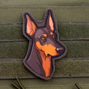 May include: A Doberman Pinscher patch with a detailed illustration of the dog's head. The patch features a brown and orange color scheme with yellow eyes and is set against a green background.