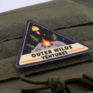 Może przedstawiać: Tr&oacute;jkątna naszywka o tematyce kosmicznej, przedstawiająca rakietę, ognisko i drzewa na tle gwiaździstego nieba. Tekst "OUTER WILDS VENTURES" jest wyświetlany poniżej. Naszywka jest przymocowana do zielonej tkaniny.