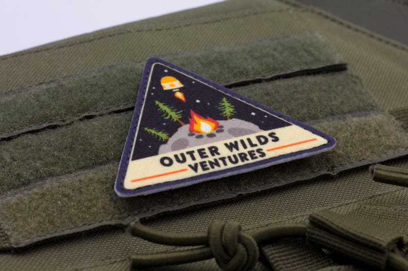 Może przedstawiać: Tr&oacute;jkątna naszywka o tematyce kosmicznej, przedstawiająca rakietę, ognisko i drzewa na tle gwiaździstego nieba. Tekst "OUTER WILDS VENTURES" jest wyświetlany poniżej. Naszywka jest przymocowana do zielonej tkaniny.