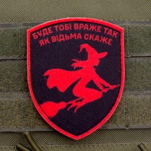 Puede incluir: Parche rojo y negro con forma de escudo, con una silueta roja de una bruja en una escoba sobre un fondo negro. Texto ucraniano visible. El parche está sobre una tela verde con cierres de velcro.