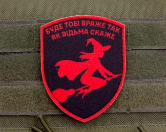 May include: A red and black patch featuring a witch on a broomstick, with text in Ukrainian. The patch is shield-shaped with a red border and is set against a green fabric background. The witch is silhouetted in red.