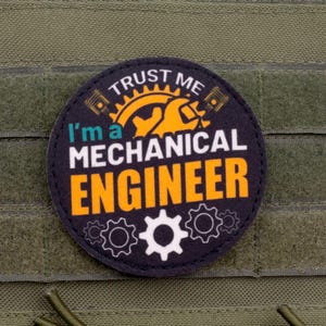 Puede incluir: Un parche redondo con el texto "Trust Me I'm a Mechanical Engineer" en letras blancas y naranjas. El parche presenta engranajes y una llave inglesa. Está adherido a una tela verde.