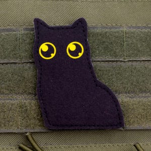 Puede incluir: Un parche negro con forma de gato y ojos amarillos está adherido a una superficie de tela verde. El parche está hecho de un material oscuro y tiene un diseño sencillo y de dibujos animados. El fondo es una tela verde oliva texturizada.