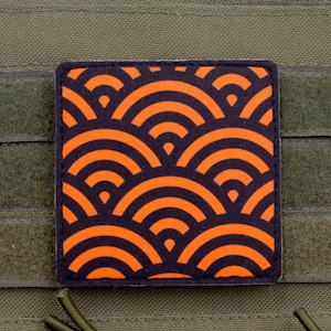 May include: A square patch featuring a repeating orange and black Seigaiha wave pattern. The patch is attached to an olive green fabric surface with horizontal webbing straps. The design consists of concentric semicircles arranged in a grid.