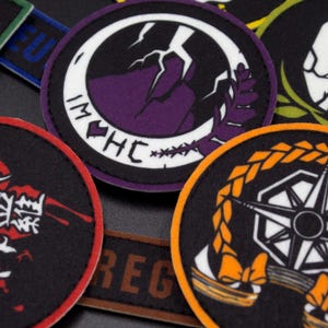 Puede incluir: Una colecci&oacute;n de parches bordados de colores con varios dise&ntilde;os, incluyendo una monta&ntilde;a morada con un rayo, una br&uacute;jula en blanco y negro y un car&aacute;cter japon&eacute;s rojo y blanco.