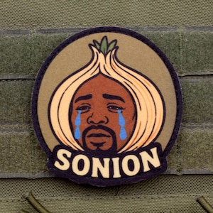 May include: A circular morale patch featuring a cartoon illustration of a person with an onion-shaped head, crying blue tears. The person has a brown complexion and a goatee. The text SONION is printed in bold white capital letters at the bottom.