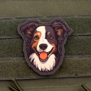 Peut inclure: Écusson coloré représentant un chien Berger Australien souriant avec une fourrure marron, blanche et noire. La langue du chien est visible. L'écusson est fixé sur un fond en tissu vert.