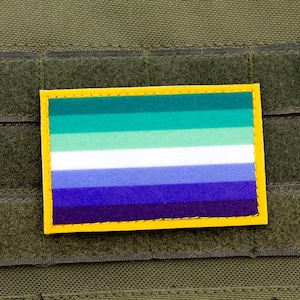May include: A rectangular patch with a yellow border and a striped design in shades of green, white, and blue. The patch is affixed to a green fabric background with hook and loop fasteners.