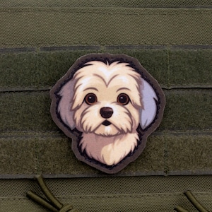 Op de afbeelding: Een patch met een cartoonillustratie van een pluizige witte hond met bruine ogen en een zwarte neus. De patch is geplaatst tegen een groene achtergrond met een klittenbandsluiting.