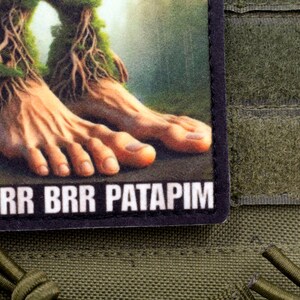 May include: A rectangular patch featuring a detailed illustration of human feet with tree-like roots and moss. The text "BRR BRR PATAPIM" is printed in white on the bottom. The patch is set against an olive green background.