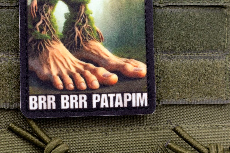 May include: A rectangular patch featuring a detailed illustration of human feet with tree-like roots and moss. The text "BRR BRR PATAPIM" is printed in white on the bottom. The patch is set against an olive green background.
