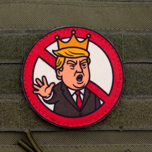 May include: A round patch featuring a cartoon image of a man with orange hair and a crown inside a red circle with a red slash. The patch is on a green fabric background.