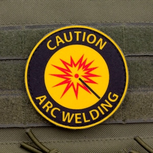 May include: A circular patch displaying the words "CAUTION ARC WELDING" in yellow on a black background. The centre has a yellow circle with a red burst and a welding torch symbol. The patch is affixed to green fabric.
