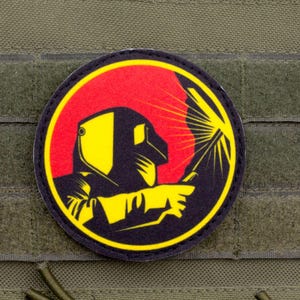 May include: Round patch featuring a welder in a yellow and black silhouette against a red background. The patch is on a green fabric with hook and loop fasteners.