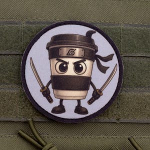 Può includere: Una toppa rotonda con un cartone animato di una tazza di caffè ninja con una fascia nera e due spade. La tazza di caffè ha un coperchio nero e una fascia nera. La toppa è su uno sfondo di tessuto verde.