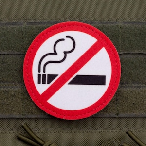 Puede incluir: Un parche redondo de prohibido fumar con borde rojo, fondo blanco y un cigarrillo negro con humo tachado por una línea diagonal roja. El parche está adherido a una tela verde.