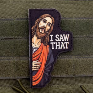 Può includere: Una toppa nera con un'illustrazione di Gesù, con lunghi capelli castani e barba, che indossa una veste rossa e arancione. Il testo "I SAW THAT" è in bianco. La toppa è su uno sfondo di tessuto verde.