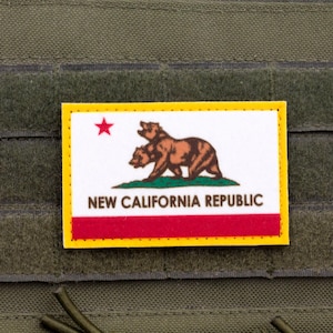 Puede incluir: Un parche rectangular con el diseño de la bandera de la New California Republic, que incluye un oso pardo, una estrella roja y el texto NEW CALIFORNIA REPUBLIC en negro, todo bordeado en amarillo sobre una superficie de tela táctica verde oliva.