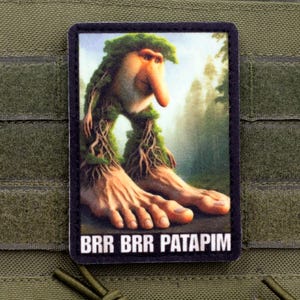 Könnte beinhalten: Rechteckiger Aufnäher mit einer skurrilen Kreatur mit langer Nase, Blätterhaaren und wurzelartigen Beinen. Die Füße der Kreatur sind groß. Der Hintergrund zeigt eine Waldszene. Der Text "BRR BRR PATAPIM" befindet sich unten.