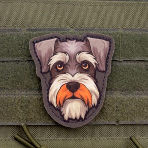 Puede incluir: Parche con la cara de un perro Schnauzer con pelaje gris y blanco, bigote naranja y ojos marrones. El parche está sobre un fondo verde con cierre de velcro.