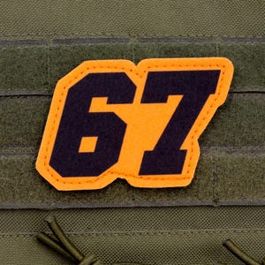 May include: A patch with the number 67 in black, outlined in orange, is affixed to a green fabric. The patch is rectangular with rounded corners. The background fabric has a textured appearance and features hook-and-loop fasteners.
