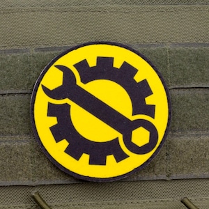 May include: A round patch featuring a black gear and wrench design over a yellow background. The patch is affixed to a green fabric surface with hook and loop fasteners, suggesting a tactical or work-related theme.