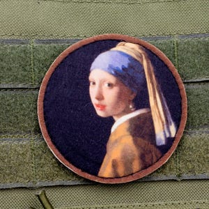 May include: A round patch featuring a portrait of a woman with a blue headscarf and pearl earring, reminiscent of the painting "Girl with a Pearl Earring." The patch is set against a green fabric background with hook and loop fasteners.