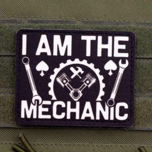 May include: A black and white patch with the text "I AM THE MECHANIC". The patch features a gear, pistons, wrench, and hammer design. The patch is rectangular and is attached to a green surface.