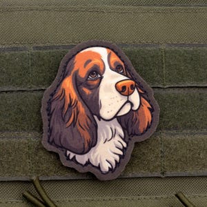 Puede incluir: Un parche con una ilustración detallada de la cabeza de un perro, que muestra pelaje marrón, blanco y negro. El perro tiene un hocico blanco y una nariz marrón. El parche está adherido a un fondo de tela verde con cierres de velcro.