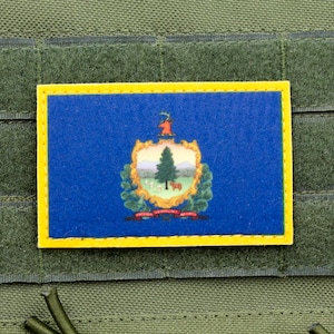 May include: A blue and yellow patch featuring the state seal of Vermont. The seal depicts a green pine tree, a mountain range, and a cow. The words "Vermont" and "1791" are visible on the seal.