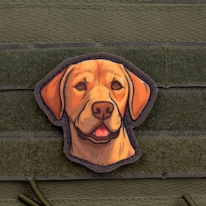 May include: A patch featuring a detailed illustration of a golden Labrador Retriever dog. The dog's head is centered, with a light brown coat, dark eyes, and a pink tongue. The patch is set against a green background.
