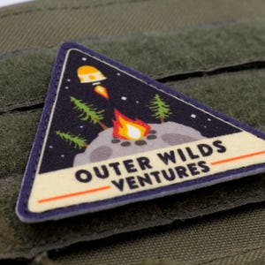 Może przedstawiać: Tr&oacute;jkątna naszywka o tematyce kosmicznej. Naszywka przedstawia rakietę, ognisko i drzewa na tle gwiaździstego nieba. Tekst "OUTER WILDS VENTURES" jest wyświetlany na białym banerze.