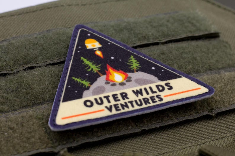 Może przedstawiać: Tr&oacute;jkątna naszywka o tematyce kosmicznej. Naszywka przedstawia rakietę, ognisko i drzewa na tle gwiaździstego nieba. Tekst "OUTER WILDS VENTURES" jest wyświetlany na białym banerze.