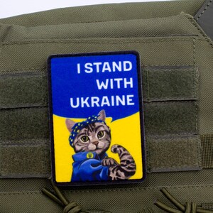 May include: A rectangular patch with the text "I STAND WITH UKRAINE" and a cartoon cat flexing its arm. The patch has a blue and yellow background, and is affixed to a green tactical vest.