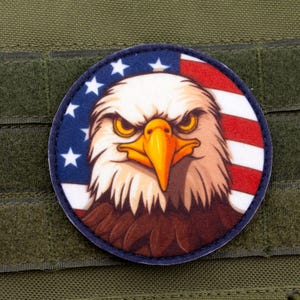 Puede incluir: Parche redondo con una ilustración detallada de la cabeza de un águila calva. El diseño incorpora la bandera estadounidense con estrellas azules y rayas rojas y blancas. El parche está sobre un fondo verde.