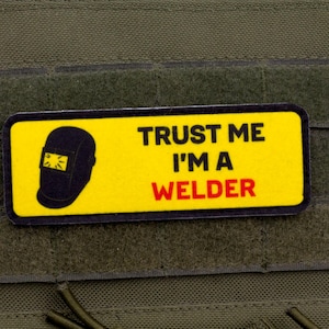 Op de afbeelding: Een gele rechthoekige patch met de tekst "TRUST ME I'M A WELDER" in zwart en rood. Een zwarte lashelm-afbeelding staat links. De patch is bevestigd aan een groene stof.