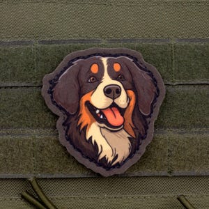 May include: A patch featuring a cartoon illustration of a Bernese Mountain Dog's head. The dog has brown, black, orange, and white fur, with a pink tongue sticking out. The patch is affixed to a green fabric background.
