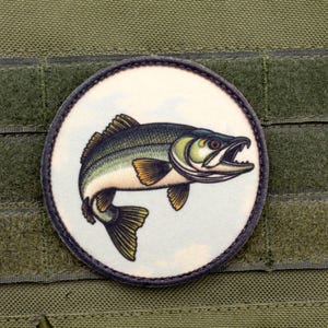 Op de afbeelding: Een ronde patch met een gedetailleerde illustratie van een vis in groen, blauw en geel. De patch heeft een donkere rand en is geplaatst tegen een witte achtergrond. Het is bevestigd aan een groene stof.